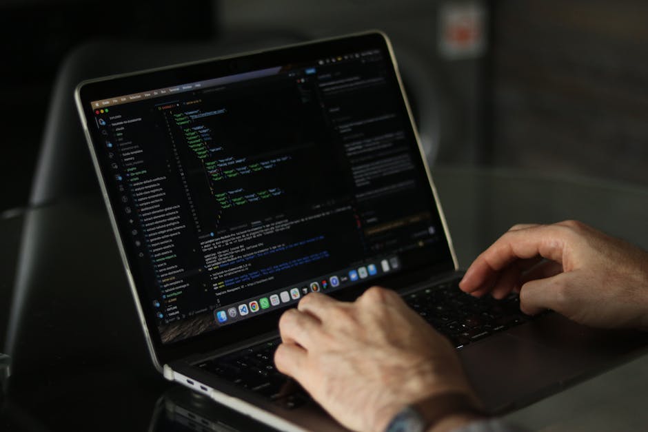 A close-up of a person's hands working on a laptop, displaying a dark-themed coding interface with lines of colorful code. The laptop is positioned on a black surface, with a blurred background sugges